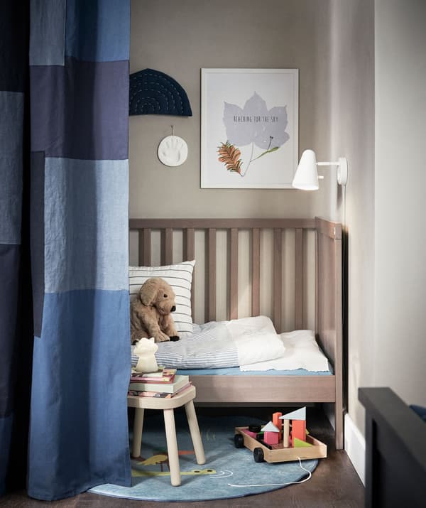 Coin d’une pièce délimité par un rideau cachant à moitié un lit d’enfant avec une lampe, des jouets, des livres et un petit chien en peluche.