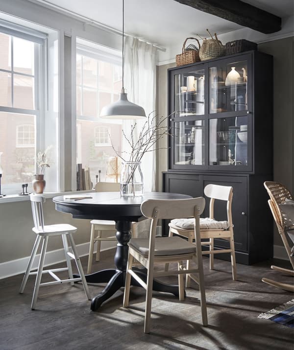 Coin salle à manger devant de grandes fenêtres. Table avec sièges pour une petite famille, à côté d’une haute armoire traditionnelle.
