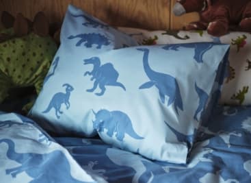 Coixí i funda nòrdica blaus amb estampat de dinosaures en un llit infantil, envoltats de peluixos.