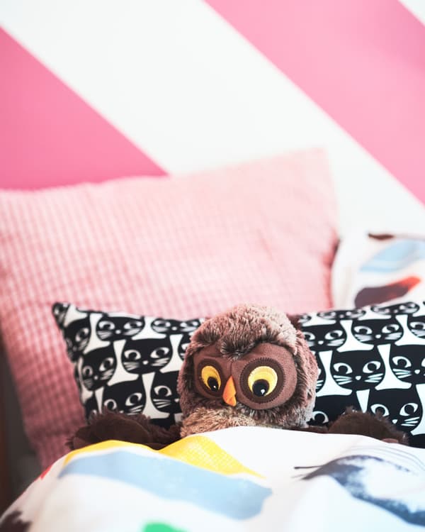 Col·lecció de coixins de color rosa suau IKEA al costat d'un altre coixí amb estampat de gats a una llitera amb un mussol de peluix.