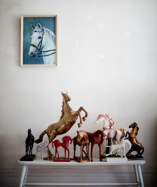 Colección de figuriñas de cabalos pousadas sobre un banco branco e cadro cun cabalo pintado colgado na parede.