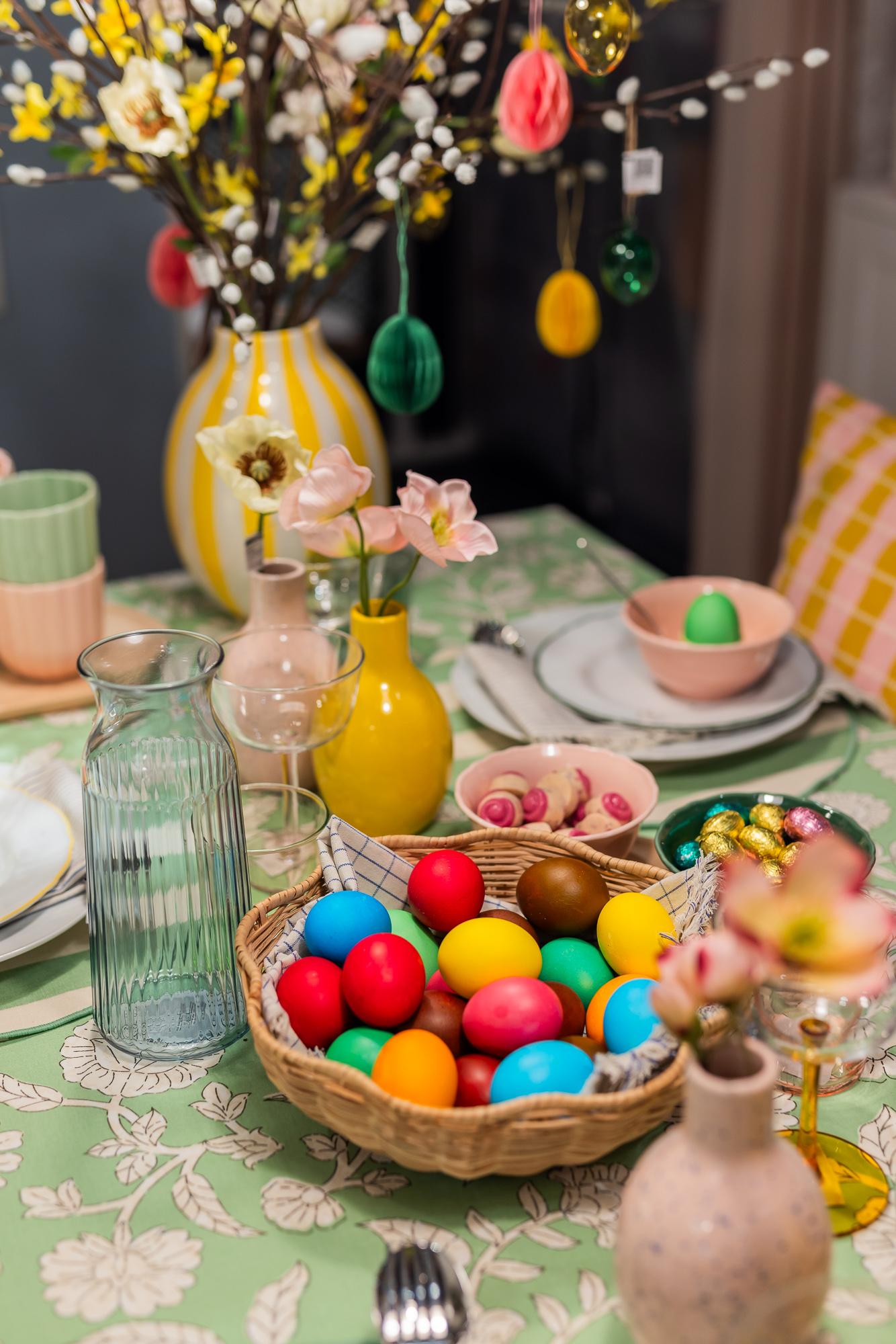Colored Easter eggs in a basket, placed in the center of the table, surrounded by vases of various shapes and colors filled with flowers and branches.