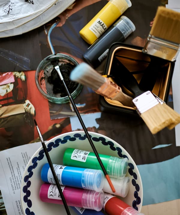 Colori a tempera in una ciotola bianca, pennelli in un barattolo nero e color oro su un tavolo coperto di giornali.