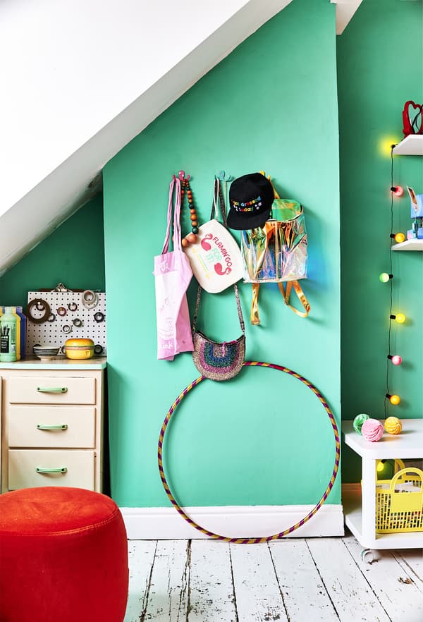 Colourful bags and a black cap hanging on hooks on a light green wall above a striped hula hoop.