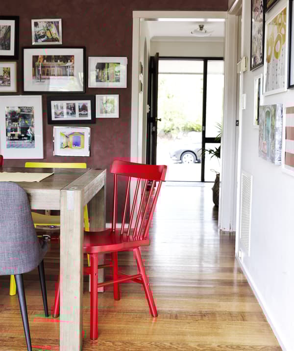 Colourful chairs around a wooden table and a dark red wall with picture gallery and a doorway into the hall.