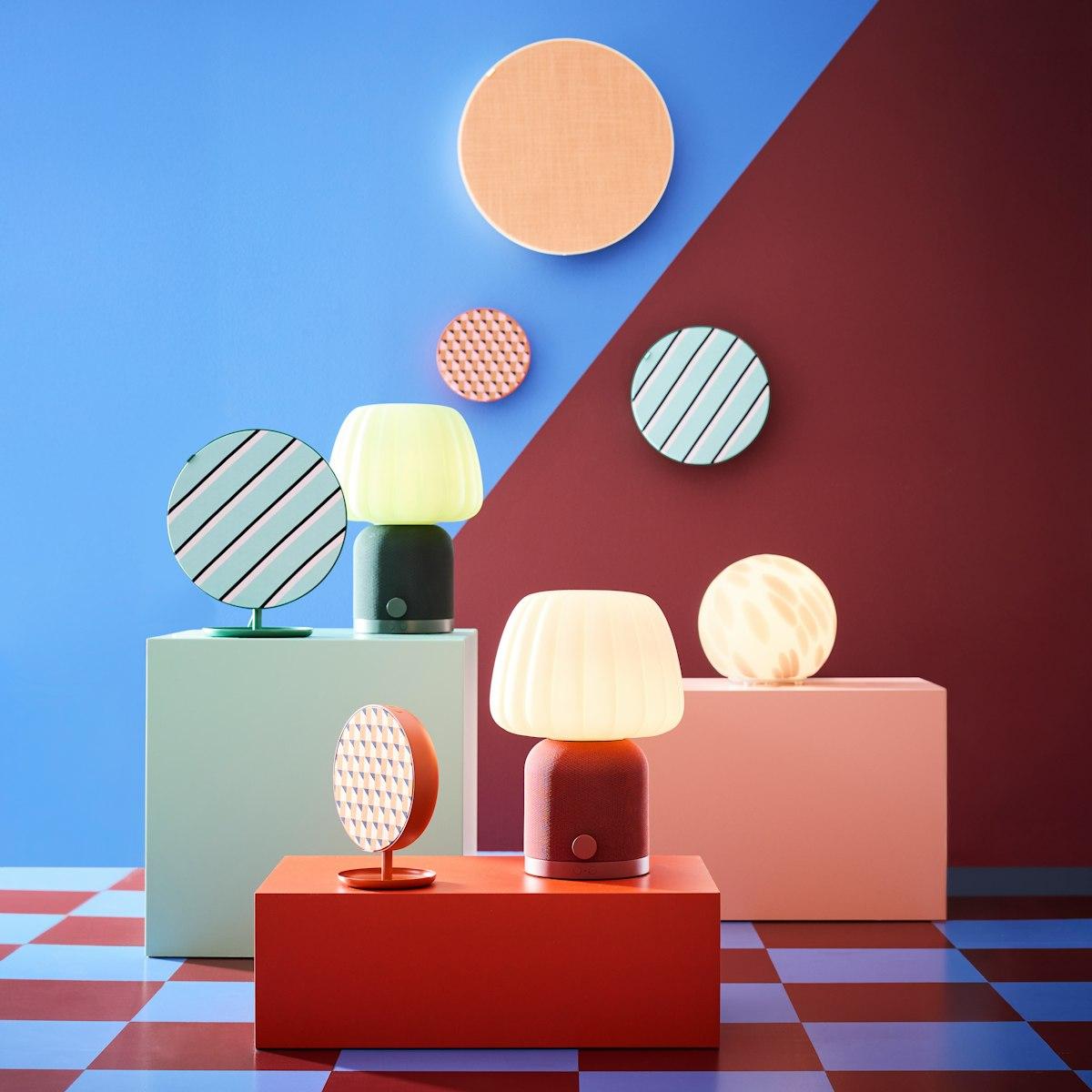 Colourful KULGLASS speaker lamps and round SOLSKYDD panel speakers arranged on blocks against a blue and burgundy geometric backdrop.