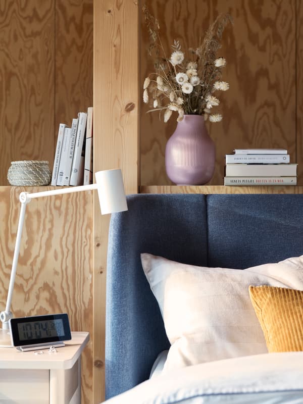 Colțul curbat al unui pat albastru TUFJORD, în fața unui perete din lemn cu o poliță încorporată, alături de o noptieră.