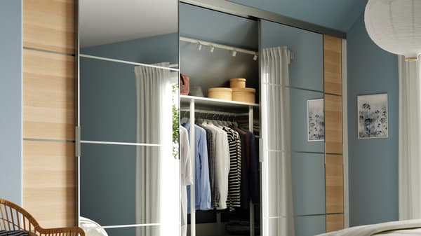 Combinaison de portes coulissantes SKYTTA dans une chambre à coucher ensoleillée. Les portes sont en partie ouvertes, révélant le rangement et les vêtements qui se trouvent derrière.