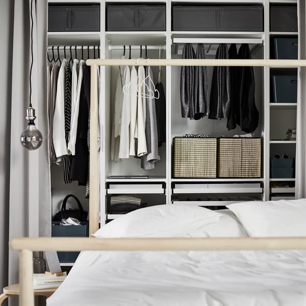 Combinație dulap PAX albă, cu mai multe secțiuni care conțin diferite haine și cutii, în spatele unui pat cu o cuvertură albă.