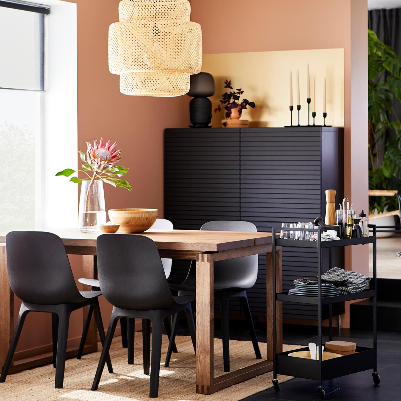 Comedor compuesto por mesa rectangular de madera con sillas color negras, junto a un carrito negro NISSAFORS. Al fondo se ve un mueble de almacenamiento BESTÅ color negro.
