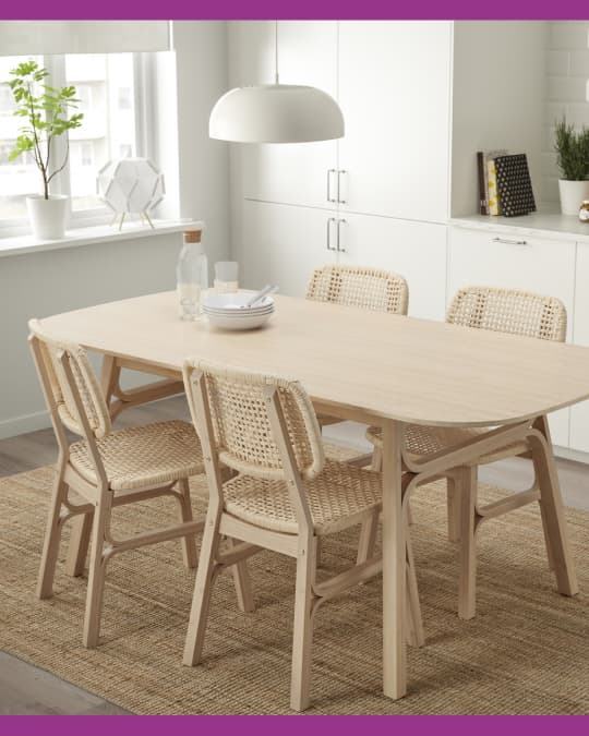 Comedor con mesa rectangular de madera clara y sillas trenzadas, acompañado de muebles blancos de almacenamiento.