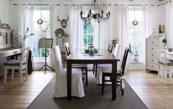 Comedor grande con decoración rústica con una mesa oscura y sillas blancas con cojines. Una alfombra grande y oscura divide el espacio.