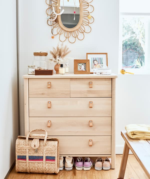 Commode en bois clair, avec miroir à cadre en osier sur le mur blanc et un sac en osier et des chaussures par terre.