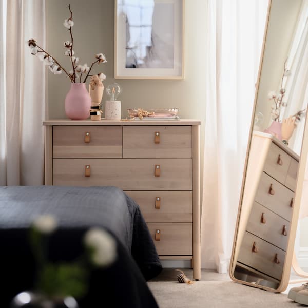 Cómoda com 5 gavetas BJÖRKSNÄS em bétula, com artigos decorativos em cima, ao lado de espelho de pé IKORNNES em freixo, num quarto.