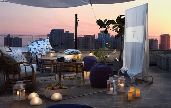 Comode sedute, tavolini, cuscini, coperte, luci a LED e uno schermo improvvisato ricreano un soggiorno sul terrazzo al tramonto - IKEA