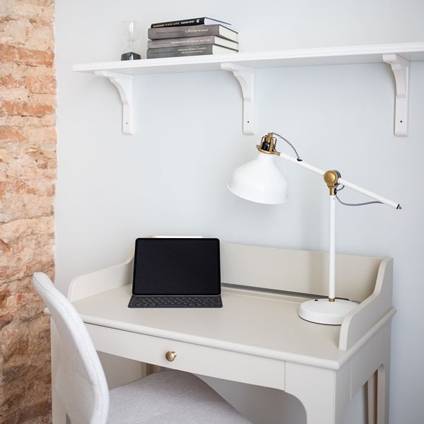 Compact and stylish home workspace featuring a small cream-colored writing desk with a brass drawer knob. On the desk sits a white adjustable reading lamp with brass accents and a tablet with a keyboard case. Above the desk, a white wall shelf holds a small stack of books and an hourglass. To the left, an exposed brick wall adds texture and contrast to the soft, light-toned interior. A cushioned light gray chair complements the clean and minimal aesthetic of the corner setup.