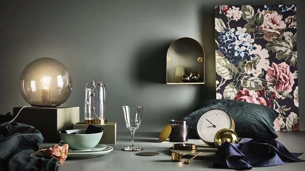 Composición de elementos decorativos sobre un fondo escuro formado por unha copa, un reloxo, unha lámpada de mesa e un fanal ou campá de vidro, entre outros.