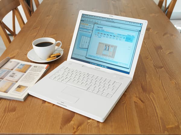 Computador portátil sobre una mesa de madera. Al costado hay un catálogo IKEA y una taza de café con galletas.