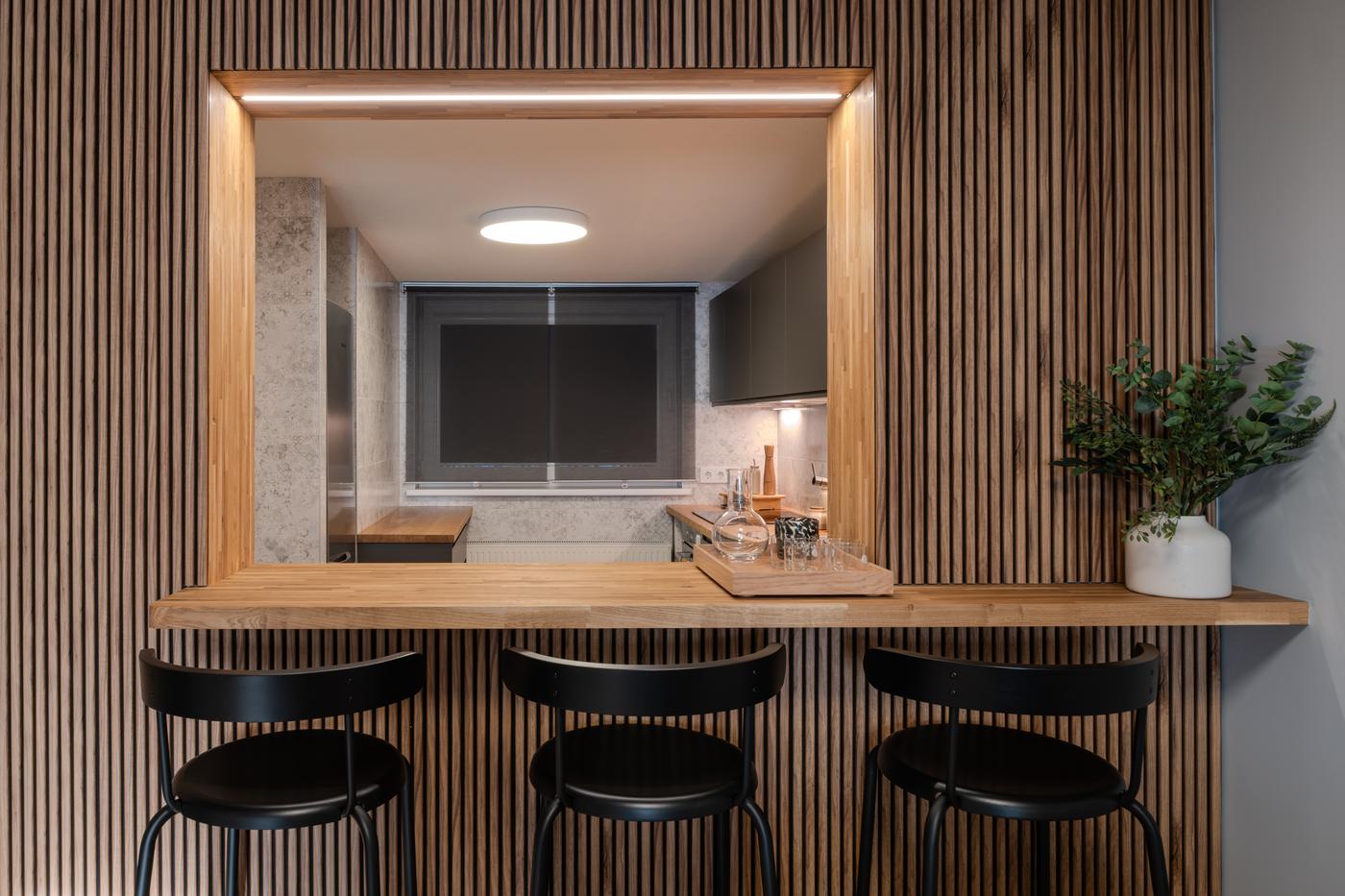 Contemporary kitchen featuring a bar zone with three black bar stools, vertical wooden panelling, and soft overhead lighting.