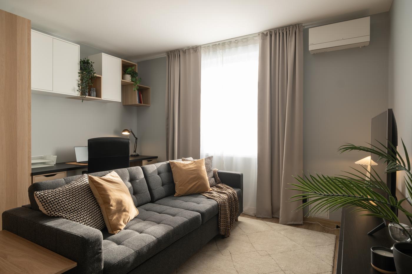 Contemporary living room with a gray sofa, light wood flooring, tall curtains, and a workspace area behind the sofa.