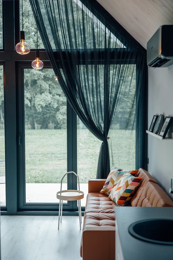 Contemporary living room with leather sofa and black sheer curtains – Stylish seating area with a tan leather sofa, geometric colorful pillows, and floor-to-ceiling windows with sheer black curtains.