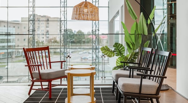 Contemporary lounge setup with red and black wooden armchairs featuring gray cushions, light wood side table, woven pendant lamp, and lush artificial plants on a black grid rug, set against a backdrop of large glass windows and urban view.