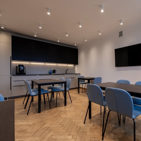 Contemporary office kitchen and dining space featuring minimalist gray and black cabinetry with under-cabinet lighting. Several rectangular dark wood tables are surrounded by upholstered light blue chairs. The room includes a coffee machine, kettle, and blue vase on the countertop. A flat-screen TV is mounted on the wall. The flooring is light herringbone wood, and the ceiling has multiple recessed spotlights.