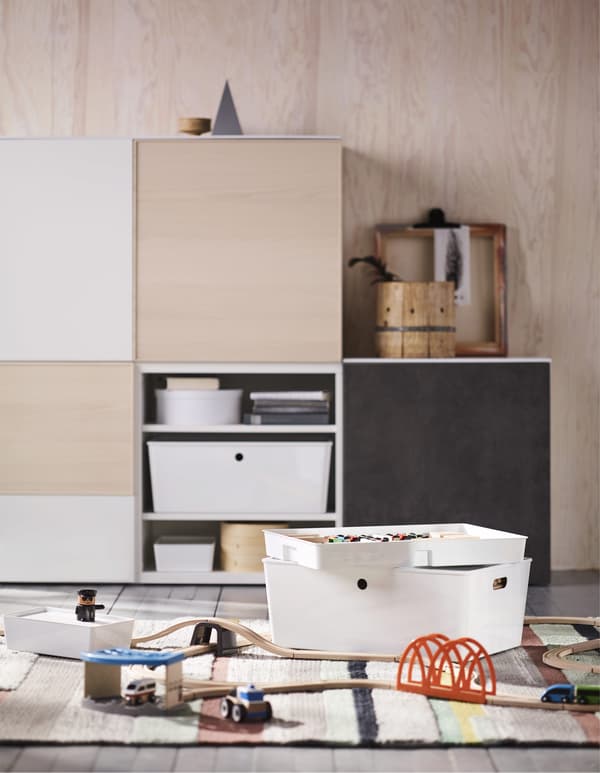 Contenitori con coperchio KUGGIS con vari oggetti come piste di legno, macchinine e un omino LILLABO su un tappeto colorato. Sulla parete di fondo, una soluzione con mobili e contenitori.