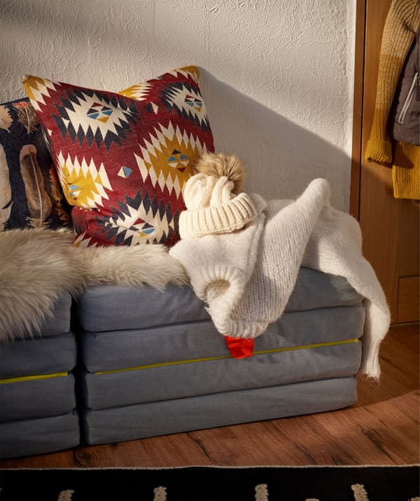 Contre le mur, un pouf/matelas replié servant de siège; coussins, peau de mouton et vêtements chauds posés dessus.