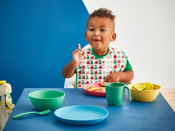 Copilul mănâncă la o masă cu vase IKEA HEROISK de culoare albastră, verde, galben și roșu din plastic PLA.