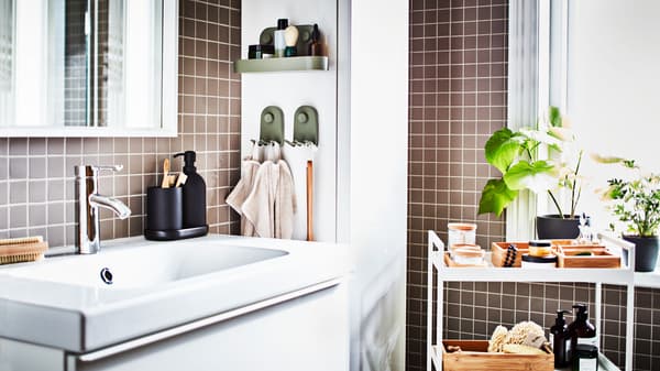 Copo para escova de dentes em cinzento escuro e doseador de sabonete colocados num lavatório em branco. Há toalhas penduradas em ganchos em verde.