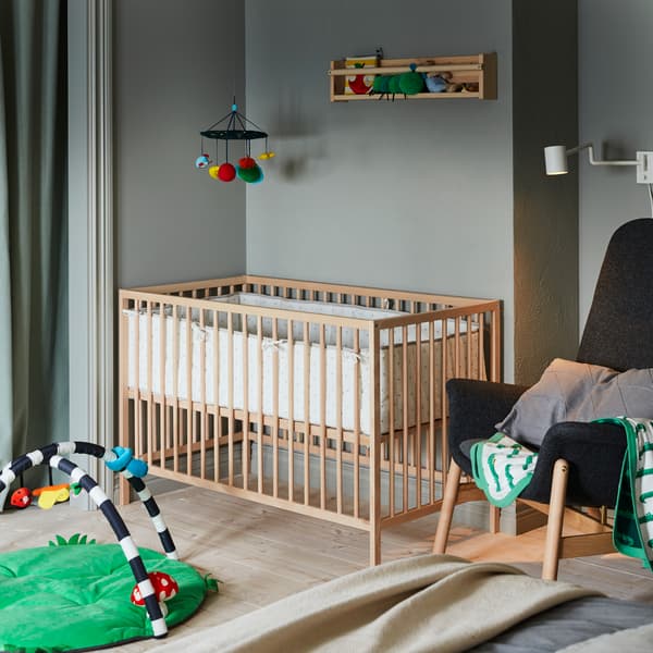 Corner of a bedroom with a wooden baby’s cot. An armchair on one side, a baby gym on another, a mobile hanging above.