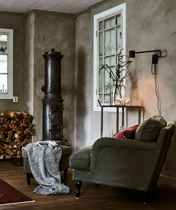 Corner of a gray-walled room with a green armchair, red cushion and footstool next to wood-burning stove and wood pile.