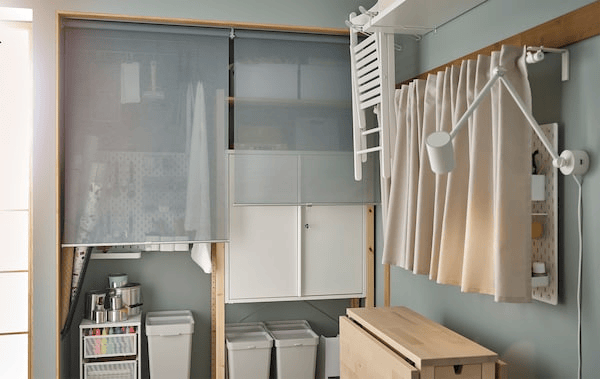 Corner of a room with hobby accessories in IVAR storage units behind roller blinds, a NORDEN table and on a draped pegboard.