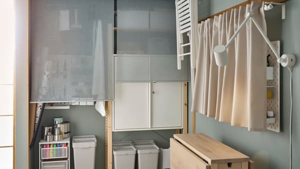 Corner of a room with hobby accessories in IVAR storage units behind roller blinds, a table and a draped pegboard.
