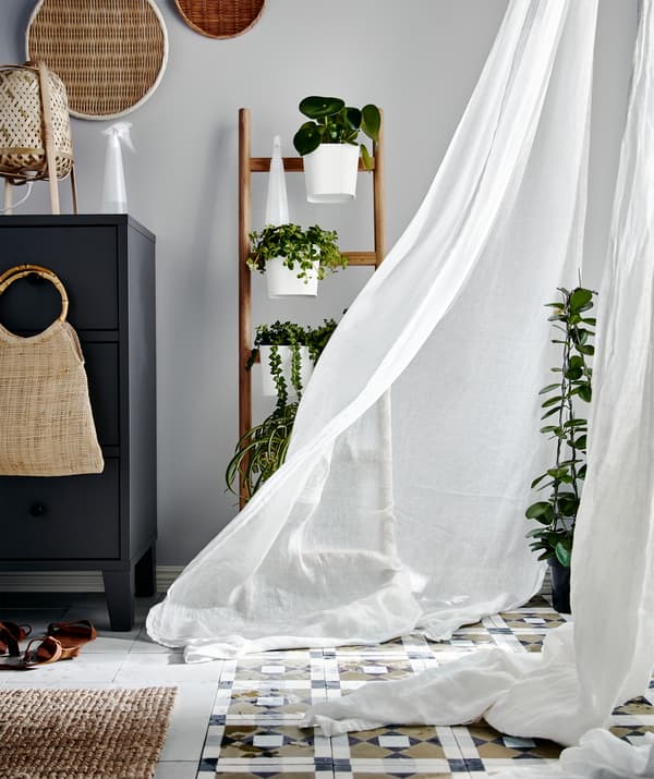 Corner of a room with plants next to a large window covered with curtains that are moving in the breeze.
