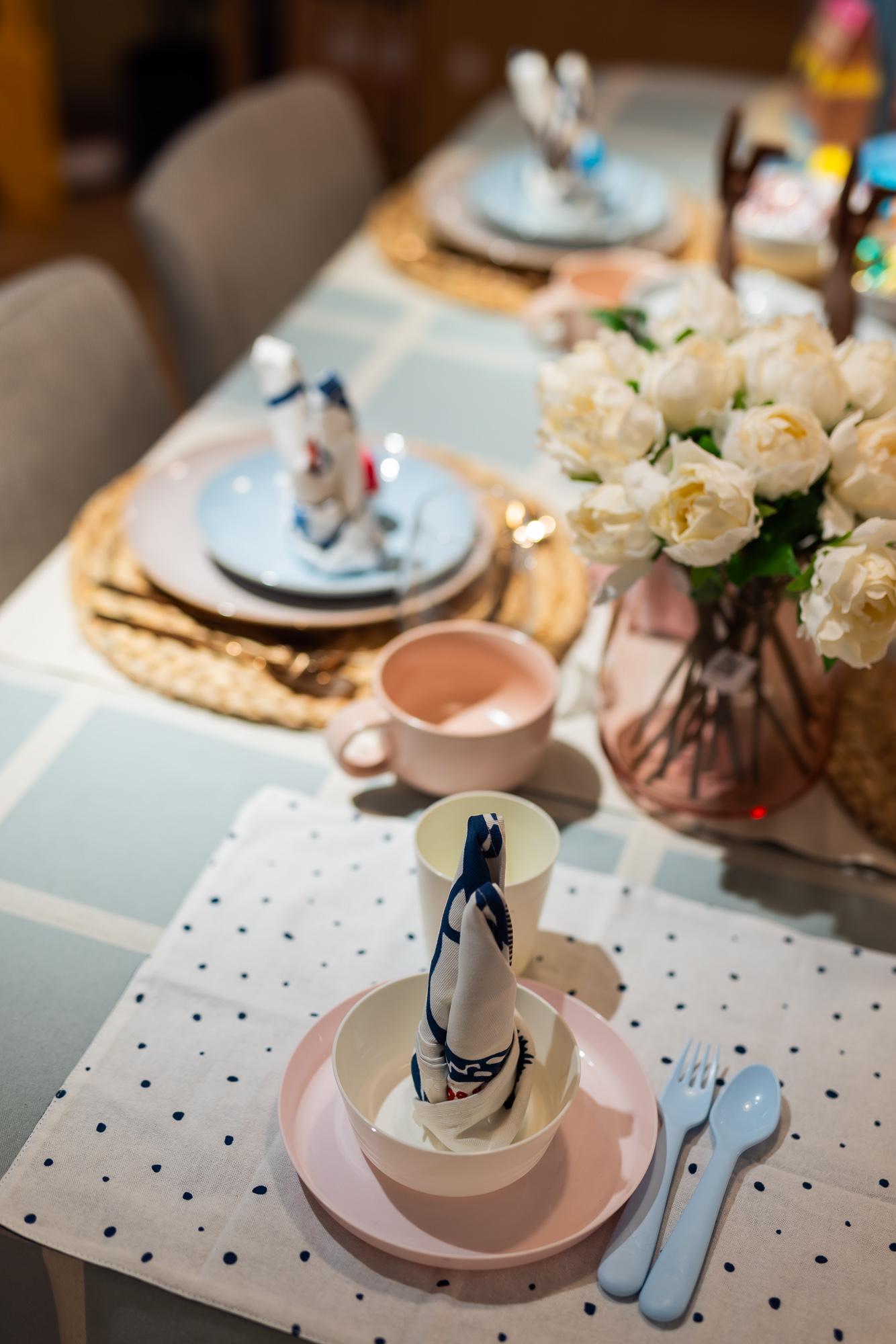 Corner of the table with a tablecloth in white and blue, and a child's seat with children's tableware in soft pink and blue.