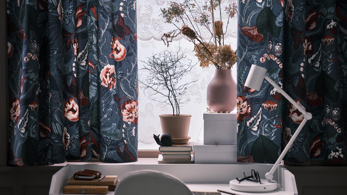 Cortinados feitos com tecido a metro FILODENDRON em azul escuro/padrão com flores, numa janela por cima de uma secretária HAUGA em branco com um candeeiro de secretária NYMÅNE em branco.