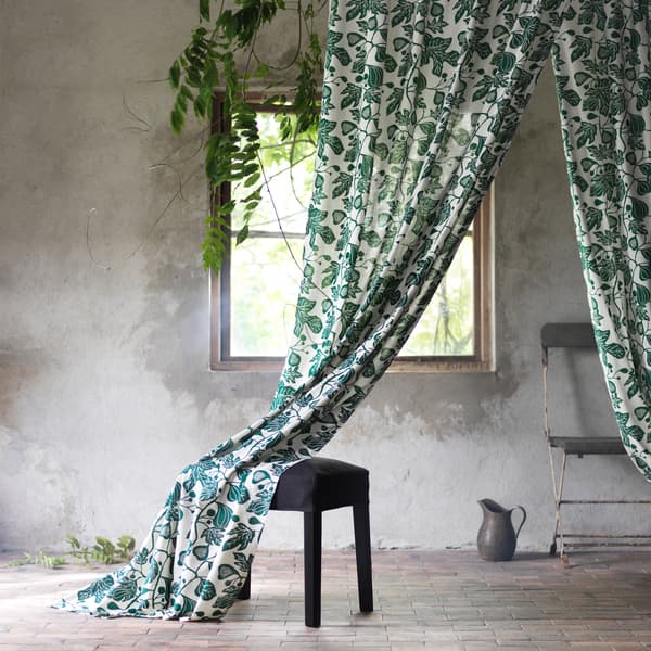 Cortinas ALPKLÖVER cun deseño de follas de figueira en branco e verde sobre un repousapés negro.