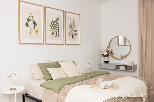 Cosy bedroom with a double bed, light textiles, decorative cushions, wall art and a round mirror above a table.