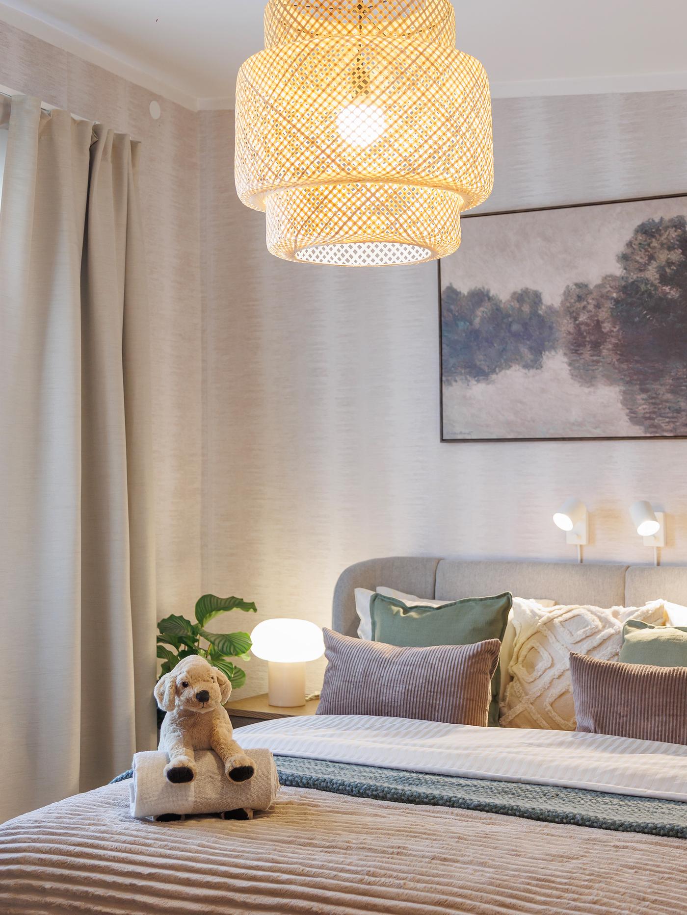 Cosy bedroom with an upholstered bed decorated with cushions, a soft toy and a woven pendant lamp above.