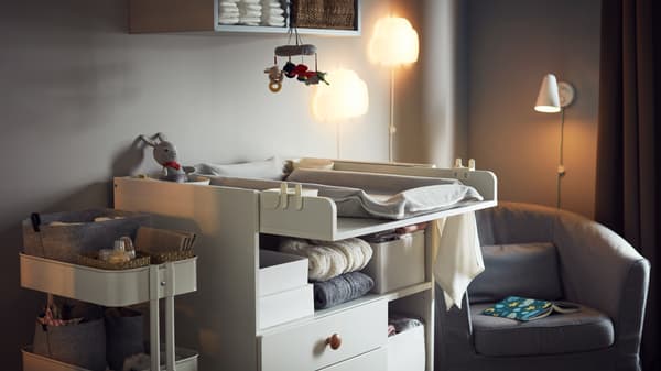 Cosy warm room with changing table, storage trolley with baby accessories, armchair, night wall lamps and wall storage.