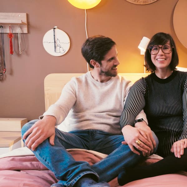 Couple assis sur un lit, l'homme brun à gauche regarde sa compagne à droite, une femme brune avec des lunettes. 