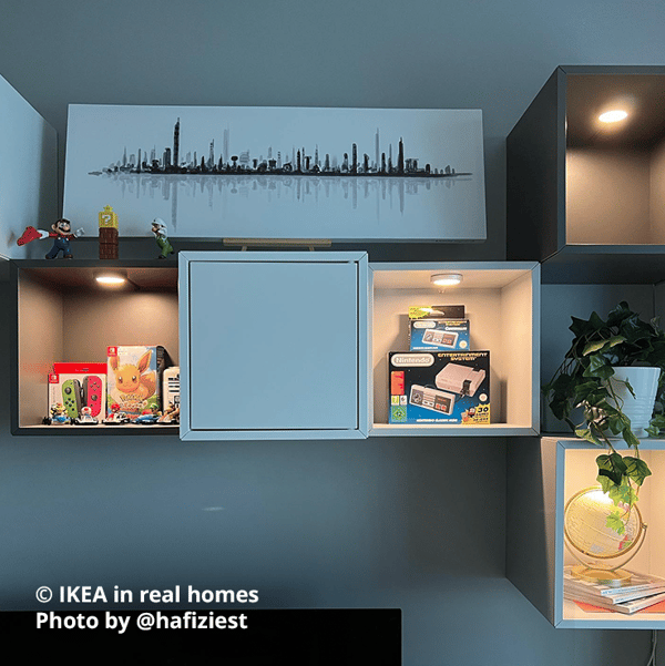 Couple of dark grey and white EKET storage cubes mounted on the wall, with MITTLED LED integrated lamps letting of a glow on the displayed items in four of them. Toys, a Nintendo Switch and game cartridges, a potted plant and some books with decorative items are displayed neatly in the cubes.  Photo by instagrammer @hafiziest for IKEA in real homes.