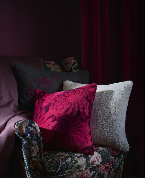 Coussins à motifs posés sur un fauteuil foncé à motif floral, sur fond rouge foncé.