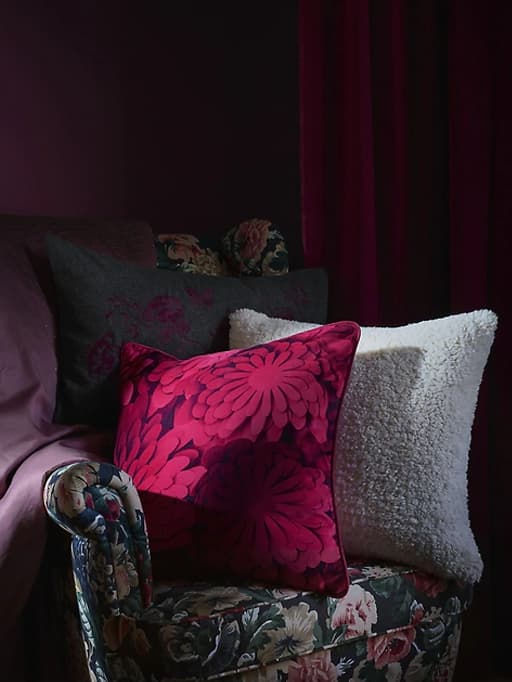 Coxíns estampados sobre unha cadeira de brazos con flores escuras fronte a un fondo vermello escuro.