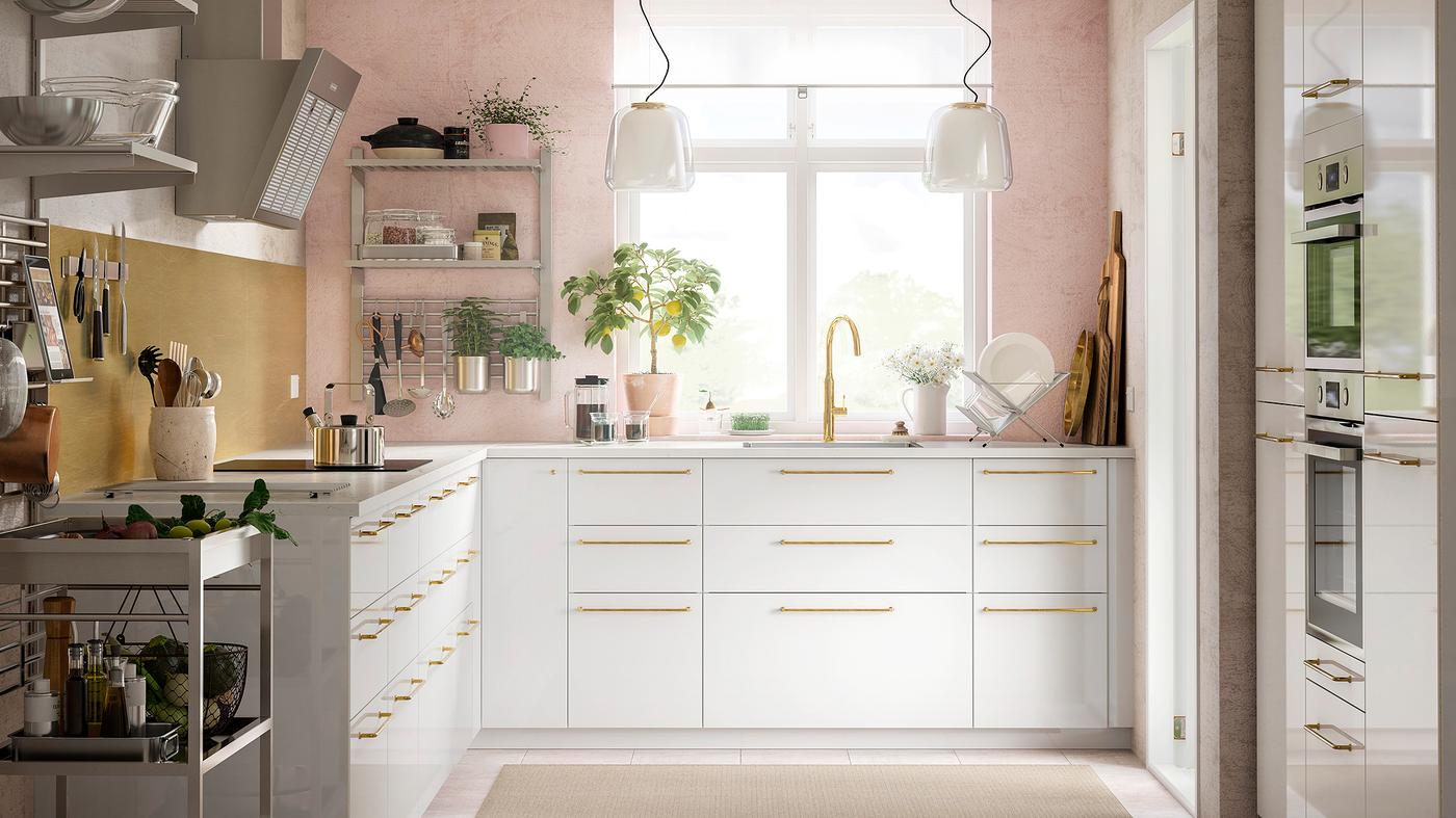 Cozinha com uma parede em rosa, com prateleiras com utensílios e gavetas e armários em branco com detalhes em cor de bronze.