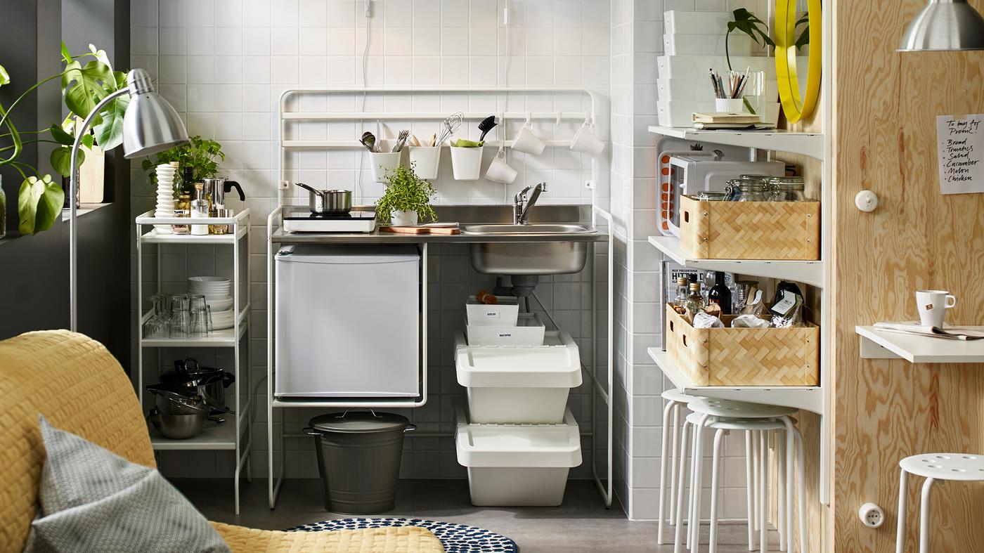 Cozinha independente em branco com uma placa de indução portátil e um pequeno frigorífico. Um carrinho de cozinha em branco onde estão arrumados consumíveis de cozinha.