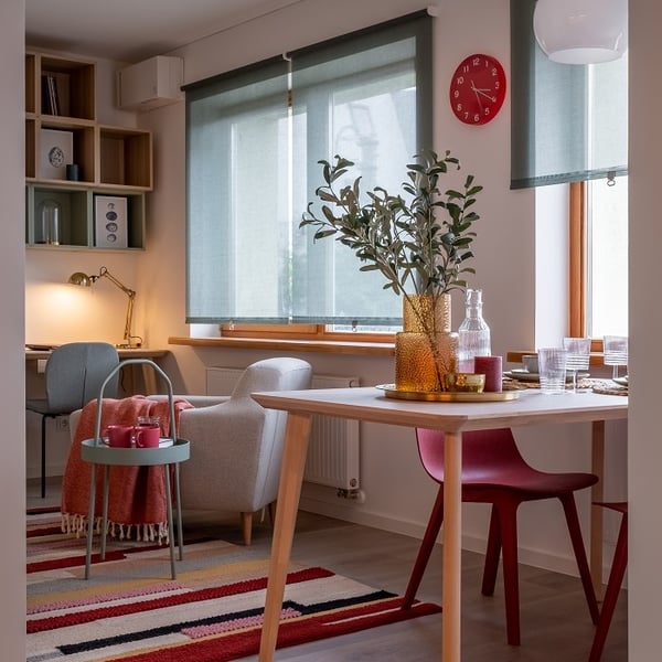 Cozy and colorful open-plan living and dining space with a modern Scandinavian design. A dining table with light wood legs is set with glassware and features a golden tray with amber glass vases and leafy branches. Nearby, a bold red dining chair adds a pop of color. The living area includes a light gray armchair, a mint green side table with red cups, and a multicolored striped rug. In the background, a small home office nook with a desk, lamp, and shelves sits under soft green roller blinds. A red wall clock adds a playful accent to the warm, inviting interior.