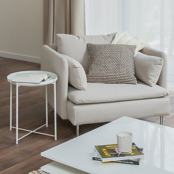 Cozy and modern living room corner featuring a light beige armchair with a black-and-white patterned pillow and a cream throw draped over the back. Next to it, a small round white side table holds an open book. In front, a glossy white coffee table displays a stacked candle and two colorful books. Floor-to-ceiling sheer curtains allow natural light to fill the space, complemented by warm wood flooring and a soft, neutral-toned palette.